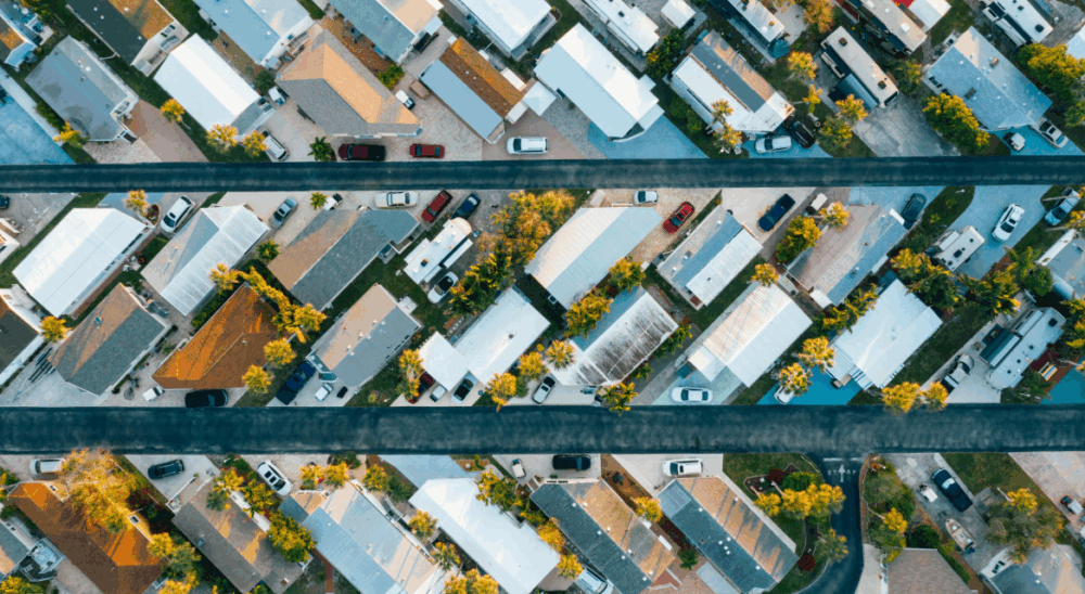 Aerial view of a suburban neighborhood with rows of houses, cars parked along two parallel streets, and visible trees in yards.